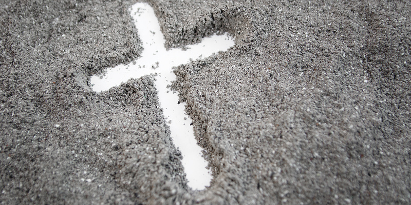 Ein mit dem Finger gezeichnetes weißes Kreuz erscheint auf grauem Asche-Hintergrund.